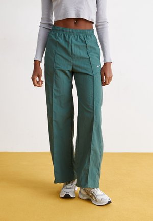 Femme portant un pantalon large Nike bleu sarcelle, un crop top blanc et des baskets blanches, debout sur un sol jaune sur fond blanc.