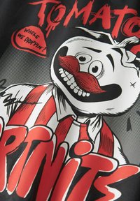 Zwarte stof met een cartoonfiguur met een witte ronde kop, een rode snor en een gestreept shirt. De tekst luidt "TOMAAI" en "WAAR GAAN WE VALLEN?" in vet rood.