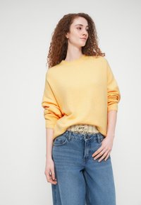 Jeune femme aux cheveux roux bouclés portant un pull jaune et un jean bleu, debout devant un fond clair uni.
