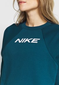 Sweat-shirt bleu sarcelle avec un col rond et des manches raglan, arborant un logo Nike blanc bien visible sur la poitrine. Tissu doux et texturé.