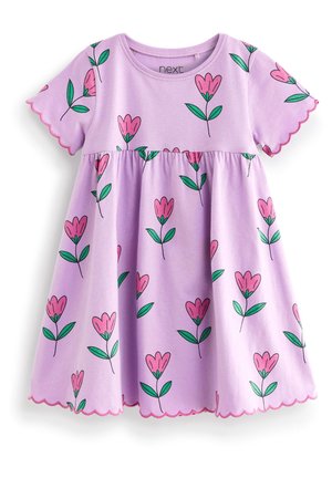 Tout-petit portant une robe violette à fleurs avec des bords festonnés et des sandales métalliques colorées, debout sur un fond clair.