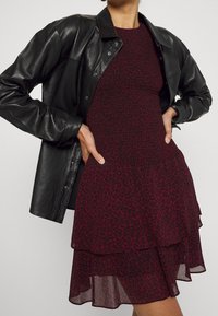 Abito con stampa leopardata rossa e gonna a strati, abbinato a una giacca camicia in pelle nera. L'outfit presenta un corpetto aderente e una lucentezza sottile.