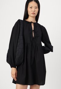 Vestido de algodão preto com um corte descontraído, apresenta mangas bufantes e um decote profundo com um detalhe amarrado. Acompanhado de uma mala de ombro preta acolchoada.