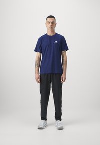 Navy blue short-sleeve athletic shirt with a subtle texture and white logo, paired with black tapered pants and light gray sneakers.