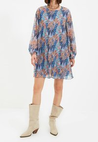 Langärmeliges Kleid mit Blumenmuster in Blau-, Rosa- und Beigetönen, locker geschnitten und mit rundem Ausschnitt, kombiniert mit beigen kniehohen Stiefeln.