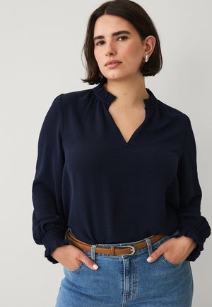 Woman with short brown hair wearing silver hoop earrings, a navy blouse with gathered collar and cuffs, tucked into blue jeans with a brown belt.