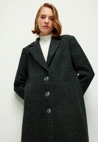 Cappotto in mistura di lana grigio scuro, con colletto a risvolto, tre grandi bottoni blu e una superficie textured. Silhouette classica e oversize.