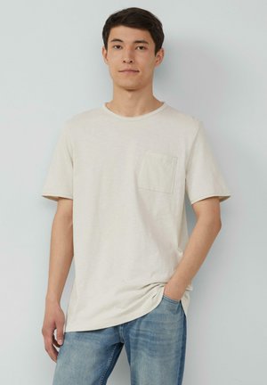 Lichtbeige t-shirt met korte mouwen en een ronde halslijn, voorzien van een borstzak, gecombineerd met blauwe spijkerbroek. Zachte textuur, casual ontwerp.
