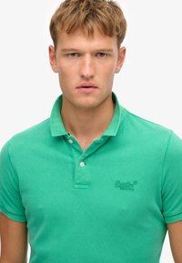 Polo verde hecho de algodón texturizado, con cuello abotonado y logotipo bordado en el pecho. Diseño ajustado con mangas cortas.