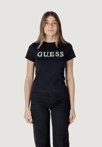 Zwarte katoenen T-shirt met witte "GUESS" logo. Korte mouwen, ronde hals, getailleerd design. Gecombineerd met zwarte broeken. Effen achtergrond.