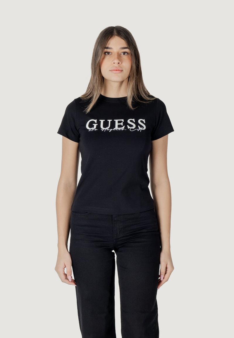 Schwarzes Baumwoll-T-Shirt mit weißem "GUESS"-Logo. Kurze Ärmel, Rundhalsausschnitt, figurbetonter Schnitt. Kombiniert mit schwarzen Hosen. Einfarbiger Hintergrund.