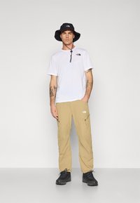 Weißes kurzärmliges Hemd mit Logo, beige Cargo-Hose mit Reißverschlüssen und schwarzer Bucket Hat, kombiniert mit schwarzen Wanderschuhen.