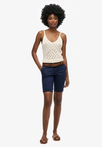 Top a canotta lavorata a maglia color crema con un motivo a diamante, abbinato a pantaloni corti blu navy e una cintura marrone. La modella indossa sandali marroni.