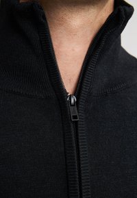 Schwarzer Reißverschluss-Pullover mit strukturierter Rippoptik und durchgehendem Reißverschluss. Nahaufnahme des Kragens und der Reißverschlussdetails.