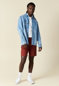 Lichtblauwe spijkerjack met borstzakken, gedragen over een witte poloshirt, gecombineerd met rode shorts en witte hoge sneakers.
