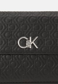 Portafoglio in pelle sintetica nera con texture, caratterizzato da un motivo stampato di 'CK' intrecciati e un'accenno di logo argentato.