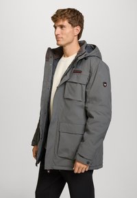 Chaqueta aislante gris con capucha, que presenta múltiples bolsillos frontales y un cierre con cremallera. Textura suave con un forro acolchado.