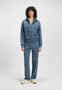 Blue tracksuit with zip-up jacket and pants in smooth, stretchy fabric. Notable logo detail on jacket and fitted design. Cream sneakers.