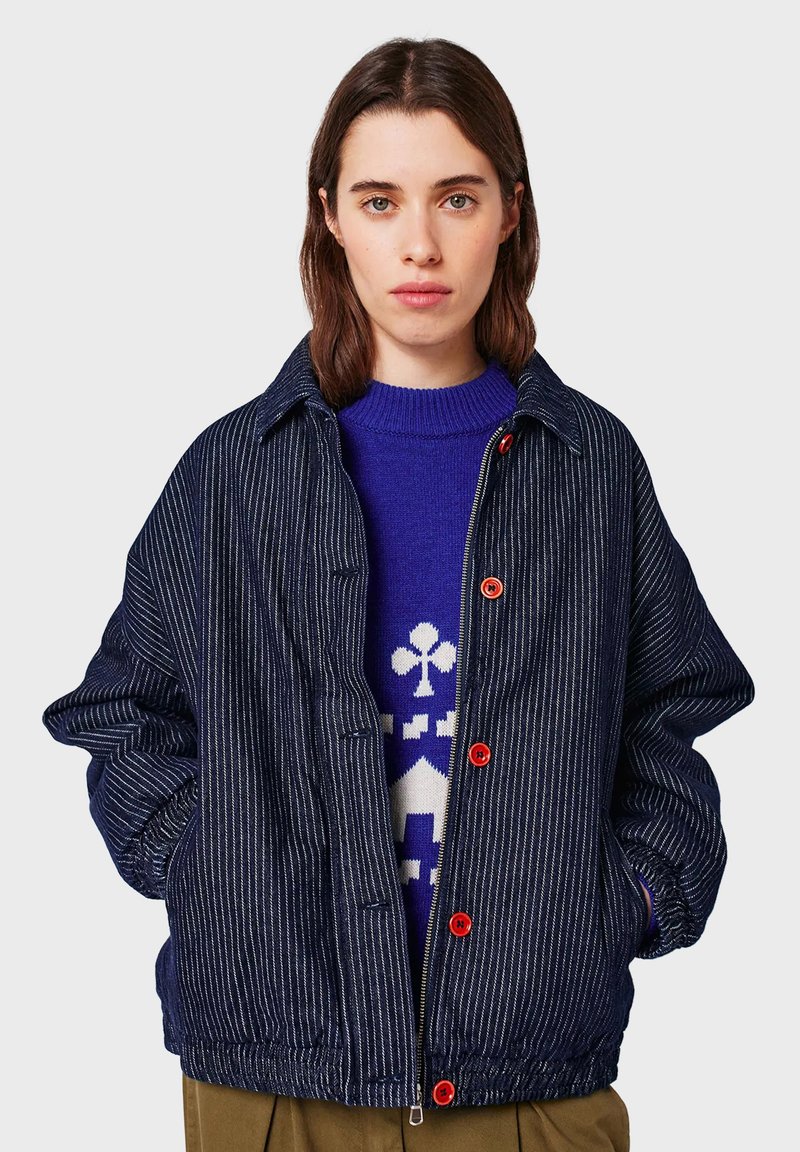 Young woman wearing a navy pinstripe jacket with red buttons over a blue sweater featuring a white geometric pattern, hands in pockets.