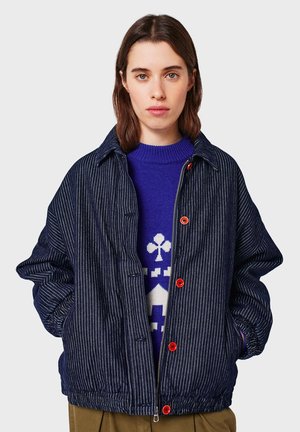 Donna dai capelli castani con indosso un giubbotto blu a righe sottili con bottoni rossi sopra un maglione a fantasia blu e bianco, con le mani nelle tasche del giubbotto.