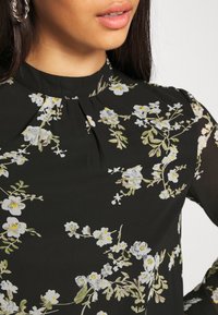 Femme portant un chemisier noir avec un imprimé floral blanc et jaune, un col montant et un détail plissé près du col.