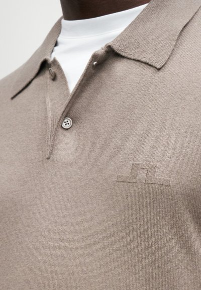 Μπεζ πλεκτό πόλο μπλουζάκι με γιακά, featuring τρία κουμπιά και ανάγλυφο λογότυπο στο στήθος. Από κάτω έχει λευκή μπλούζα με στρογγυλή λαιμόκοψη.