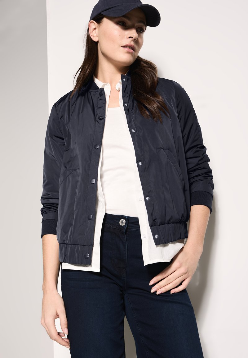 Marineblaue leichte Bomberjacke mit Druckknopfverschluss, gerippten Bündchen und Seitentaschen, getragen über einem weißen Oberteil und dunklen Jeans.