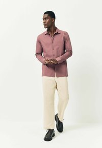 Vyras su purpuriu mygtiniu marškiniais, kreminėmis plačiomis kelnėmis ir juodais batais stovi, žiūri į šoną prieš baltą foną.