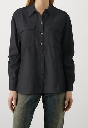 Femme portant une chemise noire boutonnée avec des poches poitrine et des manches longues associée à un jean bleu délavé.