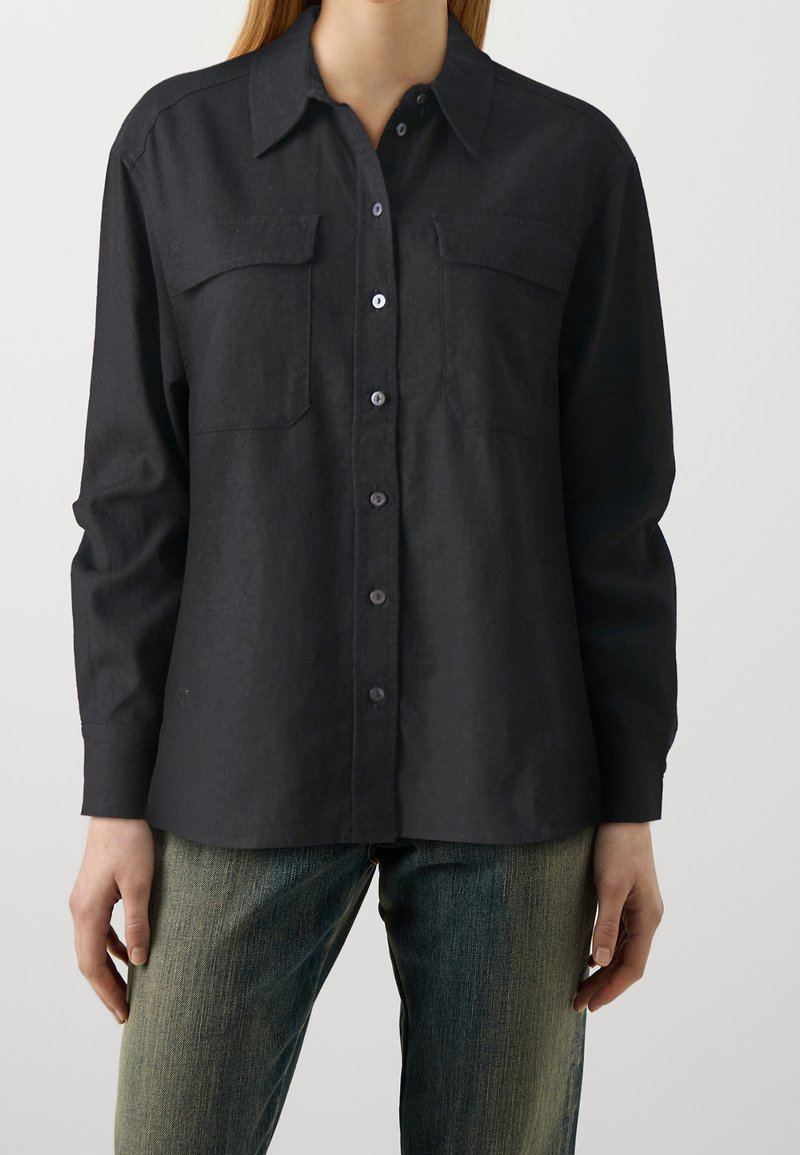 Femme portant une chemise noire boutonnée avec des poches poitrine et des manches longues associée à un jean bleu délavé.