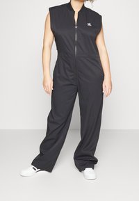 Svart ärmlös jumpsuit med dragkedja fram och elastisk midja. Har en liten vit logotypaccent på bröstet. Stylead med vita skor.