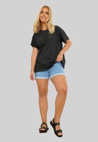 Mustat T-paita, jossa on leikattuja yksityiskohtia, yhdistettynä vaaleansinisiin farkkushortseihin, joissa on käännetyt lahkeet, sekä mustiin sandaaleihin, joissa on soljella varustetut yksityiskohdat.