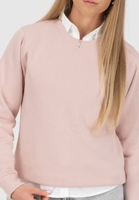 Felpa rosa chiaro con scollatura rotonda e maniche lunghe. Tessuto morbido, polsini a coste e dettaglio accentuato sul petto. Indossata sopra una camicia bianca.