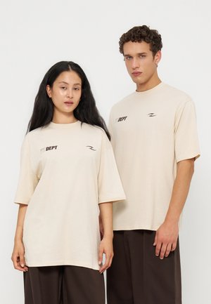 Ung mand og kvinde iført matchende beige oversized t-shirts med lille sort tekst og mørke bukser står mod hvid baggrund.