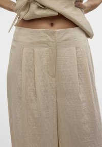Vrouw draagt losse, hoog getailleerde beige linnen broek en tilt bijpassende top op om de buik en navel te tonen, tegen een effen witte achtergrond.