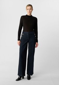 Wysokowodne spodnie z ciemnoniebieskiego denimu z ozdobami w postaci złotych guzików na kieszeniach; zestawione z dopasowaną czarną bluzką z długim rękawem i czarnymi botkami na obcasie.