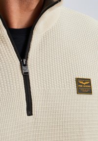 Off-white zip-up trui met een textuurpatroon, zwarte rits, en een rechthoekige merklabel in goud en zwart op de borst.