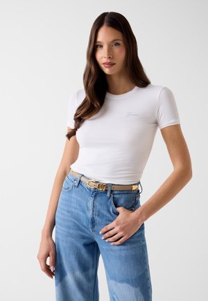 Femme aux longs cheveux bruns portant un t-shirt blanc ajusté et un jean bleu taille haute avec une ceinture beige sur un fond uni.
