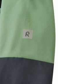 Grüne und schwarze Sportjacke mit einer glatten Stofftextur, auf der im oberen Bereich ein silbernes Logo-Patch deutlich aufgenäht ist.