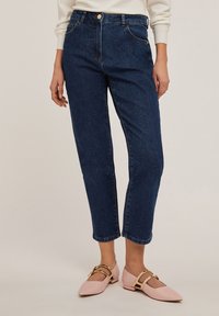 Donkerblauwe high-waisted spijkerbroek met vijf zakken, lichte stiksels en een rechte pijp; gecombineerd met lichtroze flats met gouden accenten.