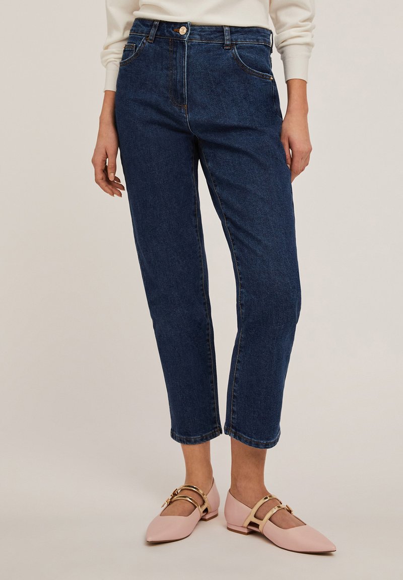 Donkerblauwe high-waisted spijkerbroek met vijf zakken, lichte stiksels en een rechte pijp; gecombineerd met lichtroze flats met gouden accenten.