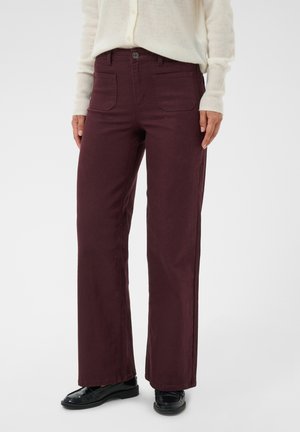 Femme portant un pantalon droit violet foncé avec poches avant, des mocassins noirs et un haut à manches longues écru avec boutons.