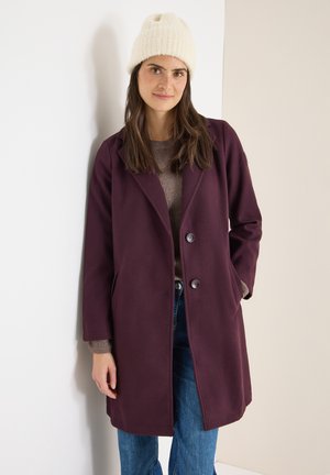 Cappotto di lana marrone con colletto, due tasche frontali e due bottoni. Indossato sopra un maglione grigio a maglia e jeans blu, abbinato a un berretto bianco.