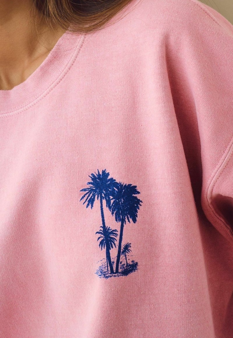Sweat-shirt rose avec un petit motif bleu de cinq palmiers sur la poitrine gauche.