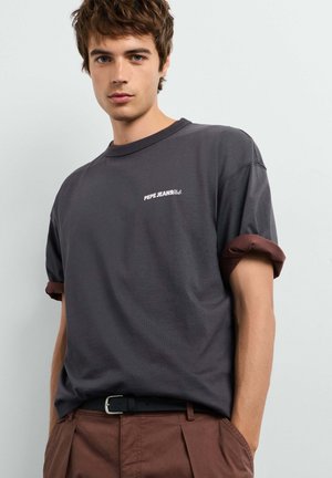 Jonge man die een donkergrijs Pepe Jeans T-shirt met opgerolde mouwen en een bruine broek draagt, staand tegen een effen lichte achtergrond.