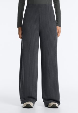 Person iført mørkegrå ribstrikkede bukser med højtaljede og vide ben, kombineret med sportsstøvler med hvide såler, stående mod en ensfarvet baggrund.