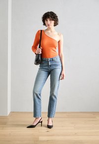 Top arancione a spalla singola, jeans azzurri a vita alta e décolleté neri con tacco. Texture lisce, design aderente e silhouette moderna.