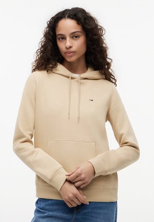 Junge Frau mit lockigem Haar, die einen beigen Hoodie mit einer Fronttasche und blaue Jeans trägt, steht vor einem weißen Hintergrund.