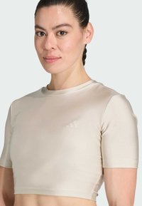 Top crop deportivo beige claro con textura suave, mangas cortas y un logo sutil en el pecho. Diseñado para actividades deportivas.