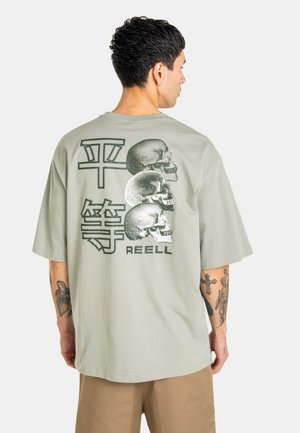 Hombre que lleva una camiseta de gran tamaño de color gris claro con tres calaveras, grandes caracteres asiáticos y el texto "REELL" impreso en la espalda.